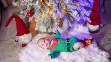 Elf yeşili kostüm, köknar ağacının altında yatan bir çocuğu giydirdi. Arka planda süslü Noel ağacı ve oyuncak cüceler. Kara kedi geliyor..