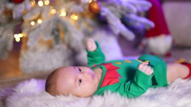 Beyaz pofuduk ekoseli yeşil cüce kostümlü küçük bir bebek. Güzel çocuk, bulanık bir şekilde Noel ağacının zemininde yatıyor..