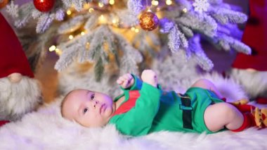 Yeşil elf kostümü giyen sevimli beyaz çocuk Noel ağacının altında yatıyor. Küçük oğlan kollarını ve ayaklarını sallıyor..