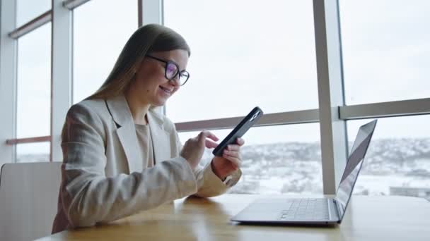 戴眼镜快乐微笑的年轻女子拨打电话号码 长头发的女士坐在靠近大窗户的书桌前 带着笔记本电脑 — 图库视频影像