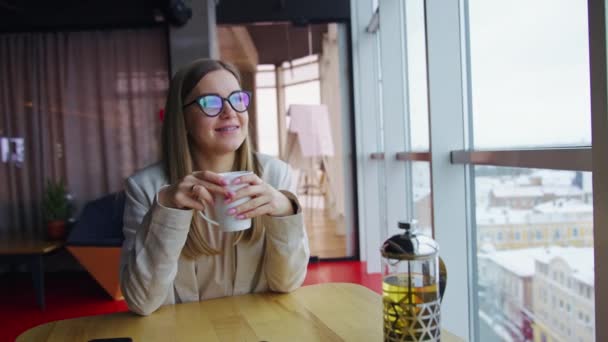 ゆったりとした女性は ランチタイムを楽しみながらお茶を飲みます テーブルに座って窓を見ている若い笑顔の女性 — ストック動画