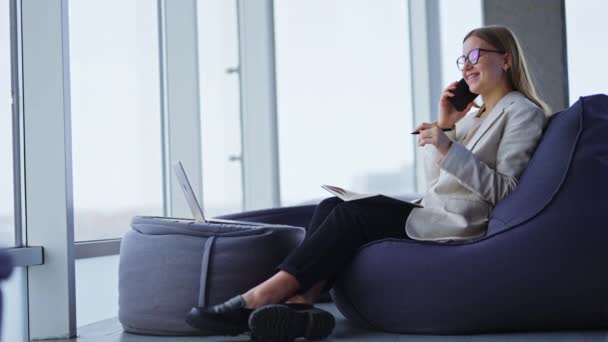 若いビジネスレディが椅子に座り 電話で話をする 左利きの女性が紙のノートにメモを書いている バックドロップでぼやけたパノラマウィンドウ — ストック動画