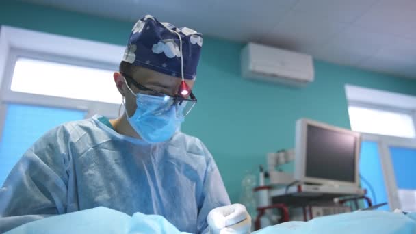 中年医生戴着蓝色的帽子 面罩和配有手电筒的器件眼镜 结束了为病人缝纫的手术 低角度视图 靠近点 — 图库视频影像