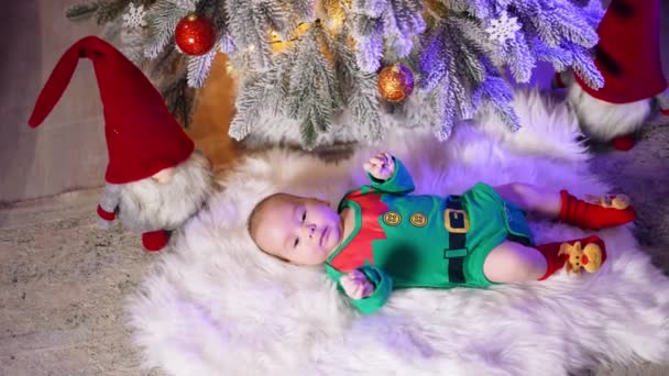 グリーンエルフのコスチュームは可愛い小さな子供を着ています かわいいトッドラーはクリスマスツリーの下に横たわる手足を動かします — ストック動画