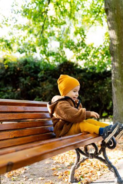 Küçük çocuk parkta bankta oturuyor ve yapraklara bakıyor..