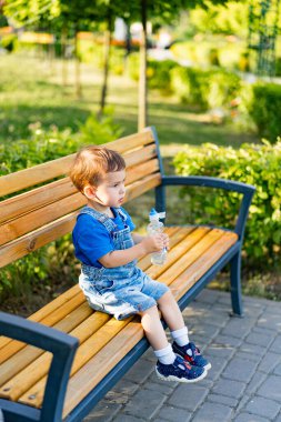 Küçük çocuk bankta oturuyor ve şişeden su içiyor..