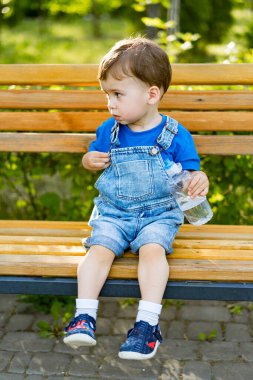 Küçük çocuk parkta bankta oturuyor ve su içiyor.