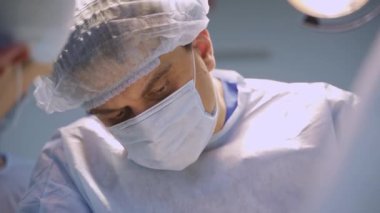 Maskeli ve şapkalı yetişkin erkek doktor ameliyat masasına bakıyor. İşbirlikçi profesyoneller ekibinde çalışan doktorlardan birinin portresi..