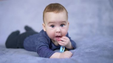 Göbeğinde mavi gömlek giyen güzel bir erkek bebek. Sakin şirin çocuk, küçücük tombul parmaklarını ağzına sokuyor. Gri arkaplan bulanık.