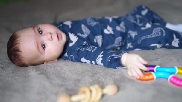 一个穿着蓝色衣服的可爱男孩躺在他的背上 可爱的婴儿把手伸向一个明亮的玩具 模糊的背景 — 图库视频影像
