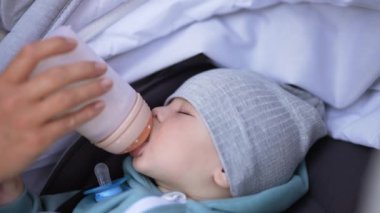 Bebek arabasında yürürken tapılası bebeği beslemek. Tatlı çocuk süt emiyor ve yavaş yavaş uykuya dalıyor. Kapat..