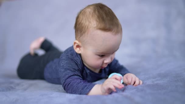 青いシャツを着たかわいい少年は腹に横たわり 手をじっと見つめている 彼の近くの太鼓でベッドの上のコーカサス人の子供 — ストック動画