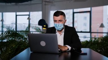 Maskeli bir iş adamı dizüstü bilgisayarın önünde oturur. Adam dikkatlice ekrana bakıyor, bir şeyler yazıyor ve konuşuyor..