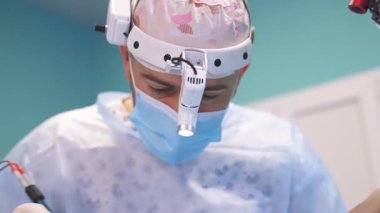 Şapkalı, maskeli ve gözlüklü orta yaşlı bir cerrah hastaya tepeden bakıyor. Cerrahlar iş yerinde alet kullanır. Kapat..