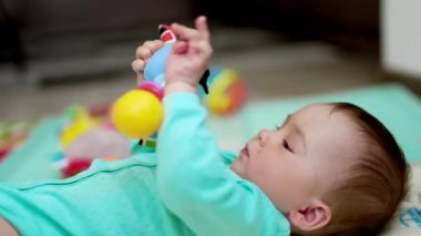 Küçük bebek bir oyuncakla oynuyor. Mavi tişörtlü şirin çocuk elinde parlak bir oyuncakla sırt üstü yatıyor. Bulanık arkaplan.