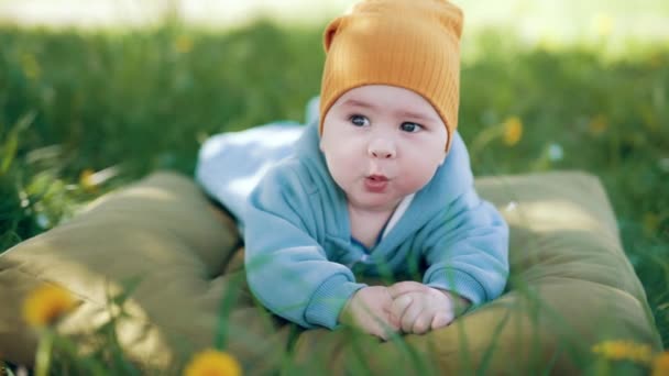 身穿蓝色衣服头戴橙色帽子的小男孩躺在绿草枕头上 可爱的孩子打呵欠 环顾四周 模糊的背景 — 图库视频影像