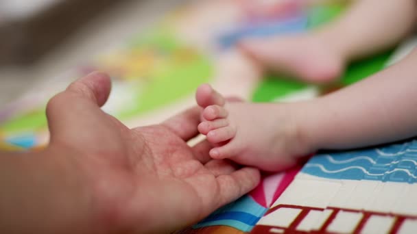 パパの手は小さな赤ちゃんの足を握っています 父親は子供の小さな指を大切にし 子供はゆっくりと指を動かしています クローズアップ ブラーレッド バックドロップ — ストック動画