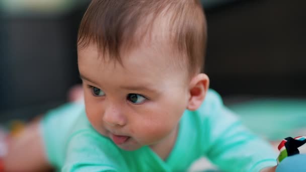 青い服を着ている親愛なる赤ん坊の少年は 腹の上に横たわっています 舌を示す素敵なアクティブな子供の肖像画 クローズアップ — ストック動画