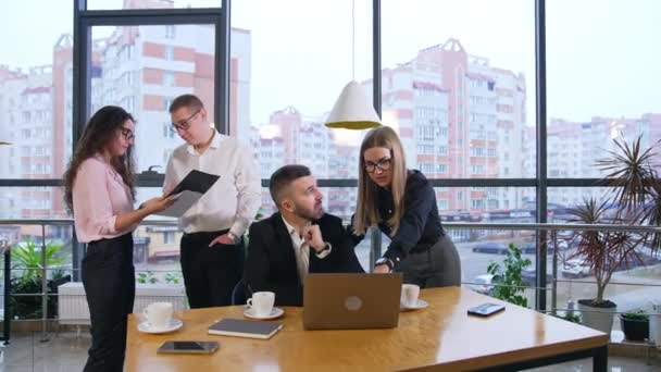 女性労働者は ノートパソコンの前に座っている上司の近くに立っている レディはコンピューターで話したり 指さしたりする 彼の腕時計を指さして急いでいるチーフ バックドロップの他の従業員 — ストック動画