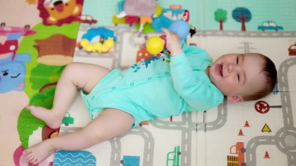 快乐活泼的白种人宝宝躺在地板上的垫子上 健康的小男孩挥动着脚和手拿着玩具 可爱的孩子在开玩笑地展示舌头 从顶部查看 — 图库视频影像