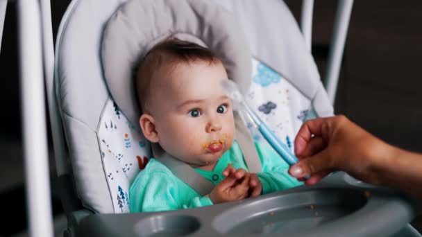 嘴脏的可爱的孩子坐在婴儿椅上 妈妈的手在给她儿子一个勺子 靠近点 — 图库视频影像