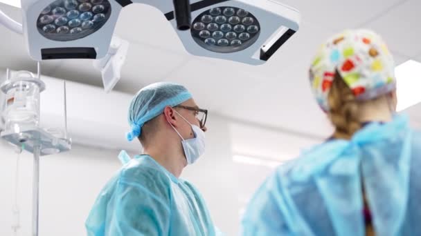 大きなランプの下に立って手術室のカウンターをドロップする男性外科医 医師はカメラの背中に立っている ローアングルビュー — ストック動画
