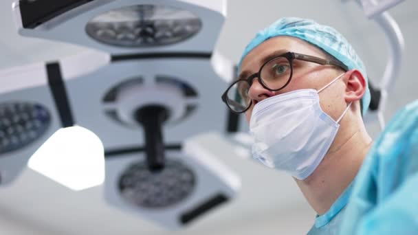 キャップ マスク メガネを身に着けている外科医の意図的な外観 彼の仕事をしている専門家の肖像画 ローアングルビュー — ストック動画