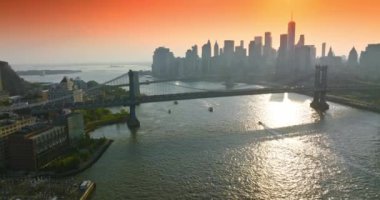 Güzel köprülerin altında East River 'dan geçen tekneler. Batan güneş ışınlarında ünlü New York gökdelenlerinin siluetleri. Pembe turuncu gökyüzü arkaplanı.