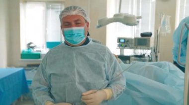 Ameliyat için gerekli donanıma sahip maskeli ve şapkalı orta yaşlı erkek doktor kamerayla konuşuyor. Cerrah, eldivenli ellerinde uzun bir iğne aleti tutuyor..