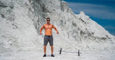 Genç atletik adam beyaz kumda halterle idman yapıyor. 