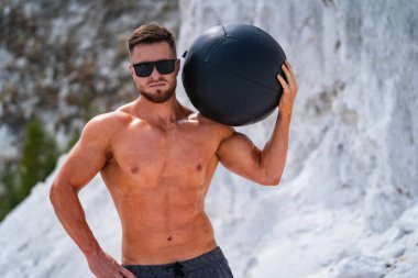 Genç atletik adam sahilde tıp topu tutuyor.