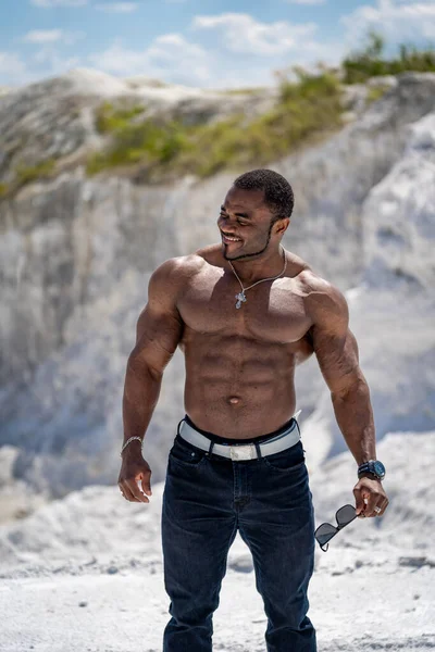 Muscular black man stands shirtless on white sand beach holding
