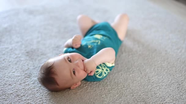 可爱的孩子把头靠在地板上对着摄像机 婴儿咀嚼着他的拳头 甜甜地笑着 模糊的背景 — 图库视频影像