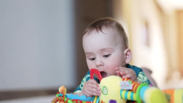 美しい赤ちゃんは おもちゃで遊んでいる床に横たわっています 子供はおもちゃから口までチューイングゴムのリングを引っ張ります クローズアップ ブラーレッド バックドロップ — ストック動画