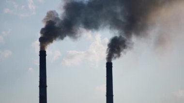 İki fabrika bacası ağır siyah duman üretiyor. Sanayi atmosferi kirletiyor. Bulutlu gökyüzü arka planı.