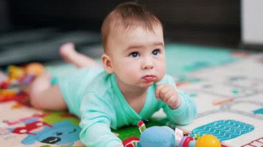 Mavi elbiseli sevimli bir bebek yerde yatıyor. Sevimli çocuk renkli bir paspasın üzerinde oyuncakların arasında yatıp etrafa bakıyor. Kapatın. Bulanık arkaplan.