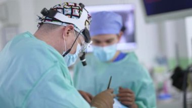 Cihaz gözlüğü, şapka ve maske takan erkek cerrah ameliyat yapar. Kameraya karşı ayakta duran doktor hastayı ameliyat etmek için araç kullanıyor..
