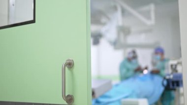 Metal saplı açık yeşil kapı ameliyathaneye açılıyor. Operasyonu bulanık bir şekilde koğuşta yapan profesyonel sağlık görevlileri.