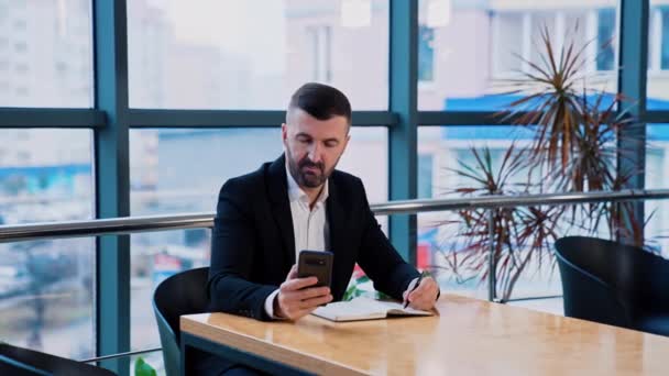 真剣に集中するビジネスマンは 机に座っているスマートフォンを使用します 電話で情報を探して 紙のノートにメモを取る男 — ストック動画