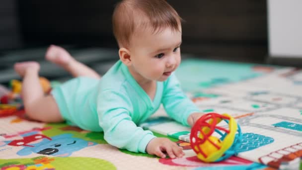好奇心旺盛なコーカサス人の赤ん坊が床に横たわってボールに達した 甘い子供はおもちゃを取って床に叩きます — ストック動画