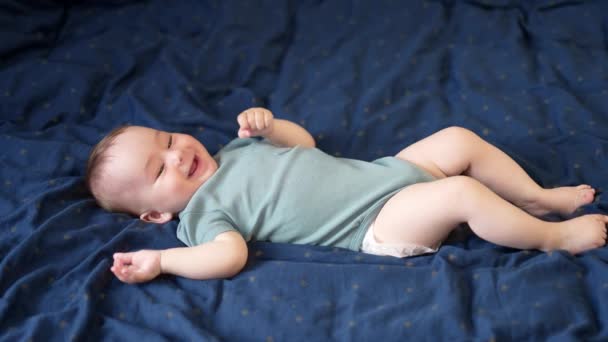快乐微笑的男婴躺在床上 可爱的孩子在他面前甜甜地笑着 蓝色背景 — 图库视频影像