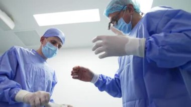 Cerrahlara ameliyat için lateks eldiven giymeleri için yardım edilir. Mavi cüppeli doktorlar ameliyata hazırlanıyor. Düşük açı görünümü.
