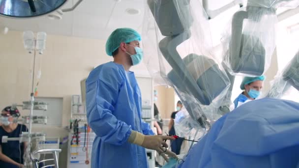 持有植入病人体内的仪器的年轻男医生 达芬奇的机器人手臂在外科医生旁边移动 低角度视图 — 图库视频影像