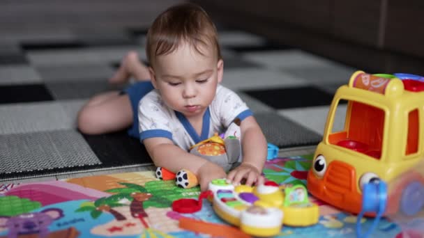 Tシャツとショートパンツの可愛い赤ん坊は 彼の腹の上のカーペットの上に横たわっています 彼の前で玩具と遊んでいる素敵な幼児 クローズアップ — ストック動画