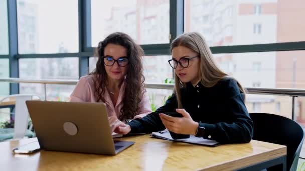 两个戴眼镜的女同事坐在办公室的桌子旁 女性在讨论工作问题时 看着面前的笔记本电脑屏幕 — 图库视频影像