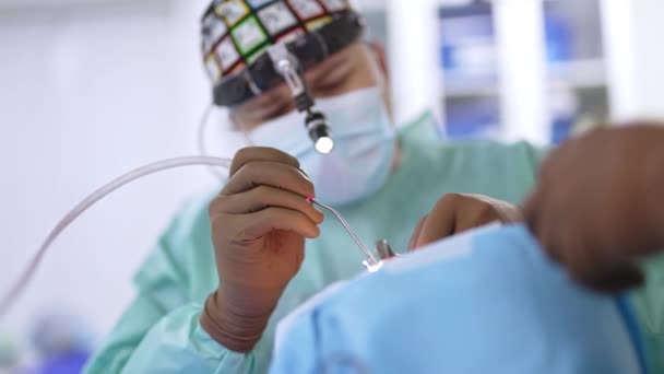 マスクの男性外科医 帽子と頭の懐中電灯は患者を操作する 医師は患者の鼻に耳鼻科の器械を適用する — ストック動画