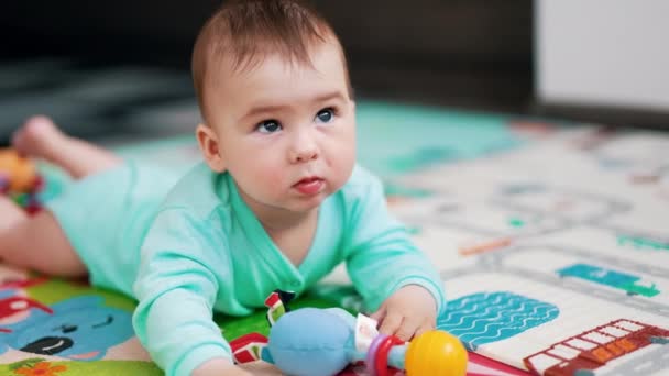 穿着蓝色保险杠的可爱的小男孩躺在彩色的垫子上 可爱的孩子玩玩具 把它拖到嘴边 饶有兴趣地看着它 — 图库视频影像