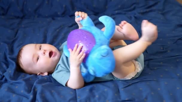 かわいいコーカサス トッドラーは背中の柔らかいベッドの上に横たわっている 赤ん坊の少年はそれと遊ぶ彼の腹の上に大きいおもちゃの亀を持っています サイドビュー — ストック動画