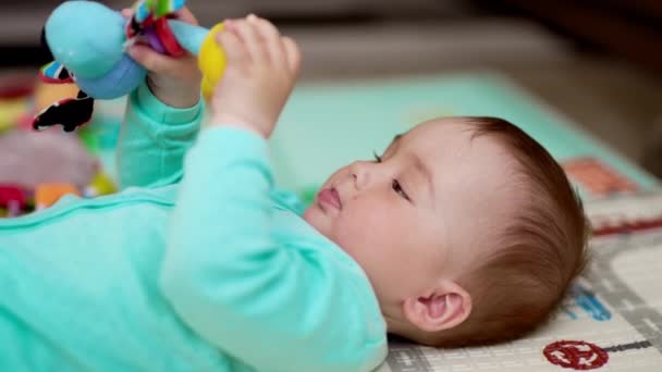 穿着蓝色衣服的可敬的健康的白人小孩躺在他的背上 活泼的婴儿在空中挥动着他的玩具 靠近点模糊的背景 — 图库视频影像