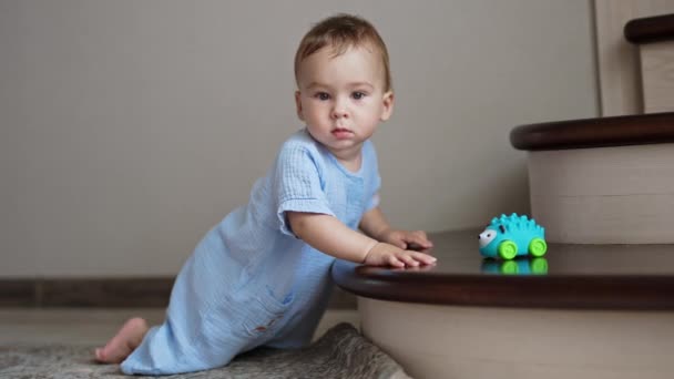 和平的白人男婴穿着蓝色保险杠站在楼梯旁边 一个玩具躺在楼梯上 但小孩却把它放在一边看 — 图库视频影像
